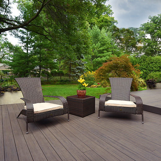 Two dark brown chairs and a brown accent table with a reddish vase are on top of a dark brown deck adorn with Espresso decking from the Legacy Collection. Varying shades of green trees appear in the background.