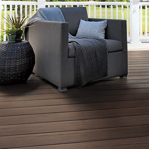 A dark gray armchair with a gray throw and light blue pillow next to a dark wicker planter on a warm brown composite deck in Dark Cocoa from Prime + Collection.