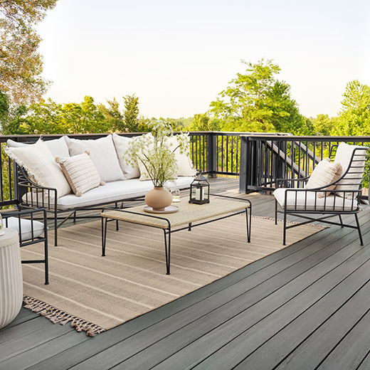 A black wrought iron couch with white pillows, a light brown coffee table, and a light brown rug atop gray decking in Sea Salt Gray from Prime + Collection, trimmed with black railing.