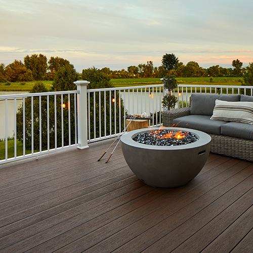 A concrete fire pit with low orange flame on a patio with white railing and gray furniture decorated with dark brown decking called Dark Roast from the Reserve collection.