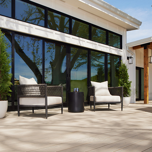 Dark brown furniture with white pillows on a bright patio with a round black end table on very light beige decking in Reclaimed Chestnut from the Reserve Collection. The chairs sit against a wall of windows reflecting a big mature tree.