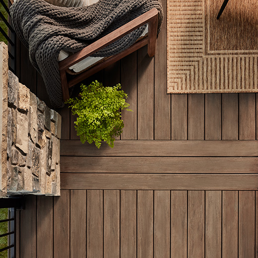 An overhead view of brown decking in English Walnut from the Vintage Collection. There is a portion warm brown arm chair with a dark gray throw next to a cobbled walk and tan woven rug.