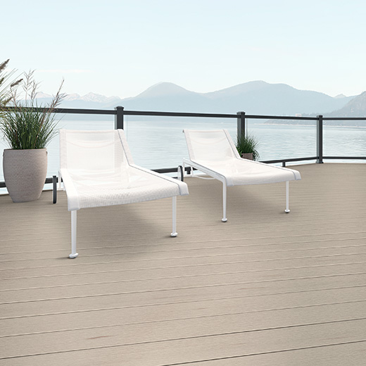 Two white chairs sit atop beige decking known as Weathered Oak from the Terrain+ Collection. A silhouette of mountains are in the back image and light colored planter sits next to the white chairs. The railing is black and see through.