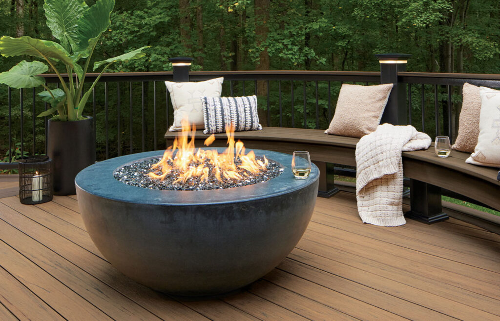 A round fire pit on a wooden deck with a curved bench and pillows, surrounded by a forest background. Two glasses of wine sit on the table and bench.