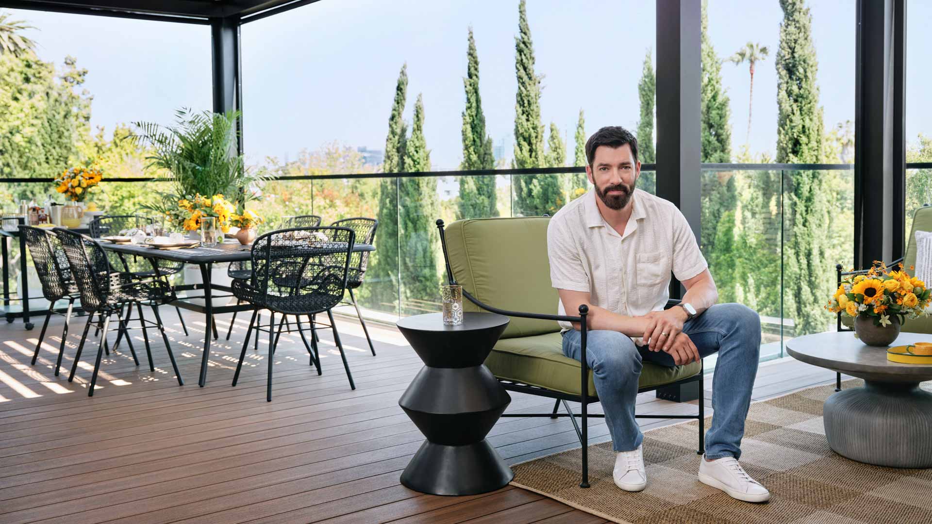 Drew Scott sits in a green chair on his deck, a table and chairs are in the background