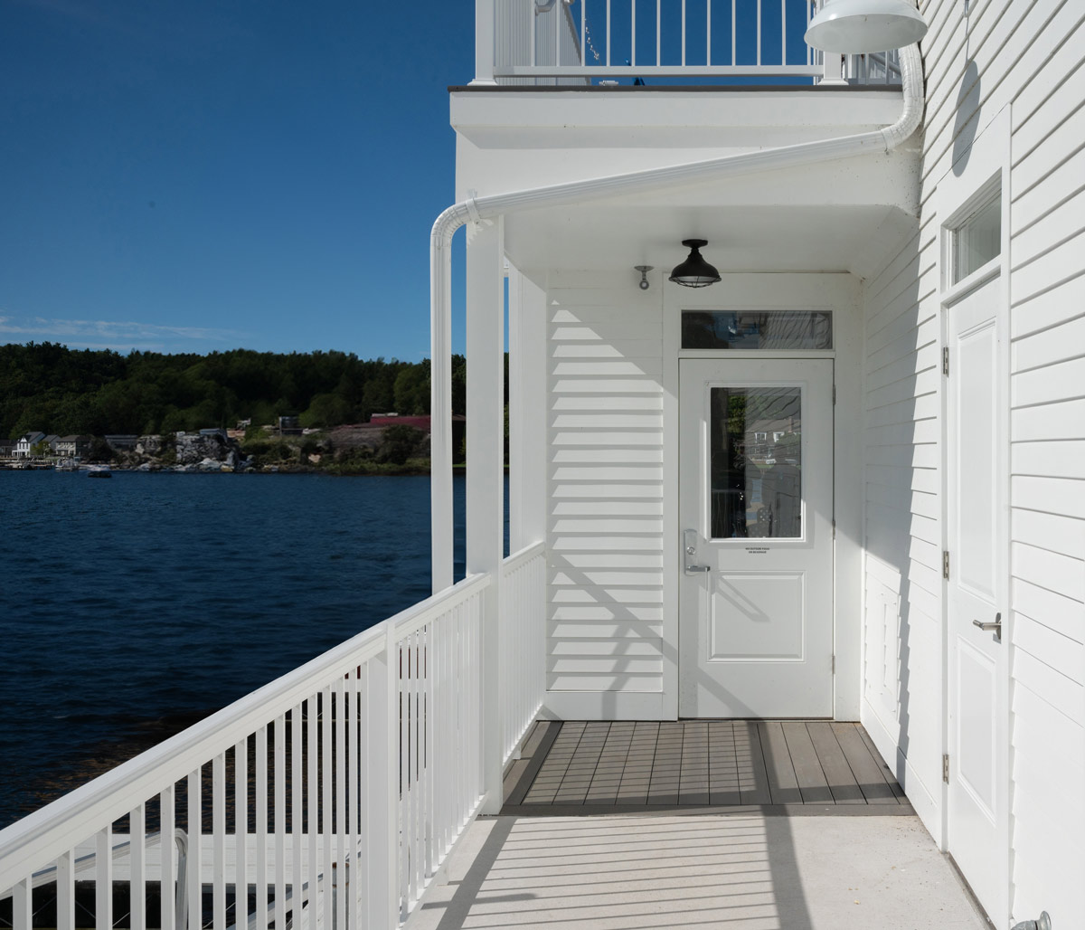 A closer view of the white railing o the yacht club with dark blue water in the background and light blue sky above with a dark multicolored divider made of residences and local businesses. 