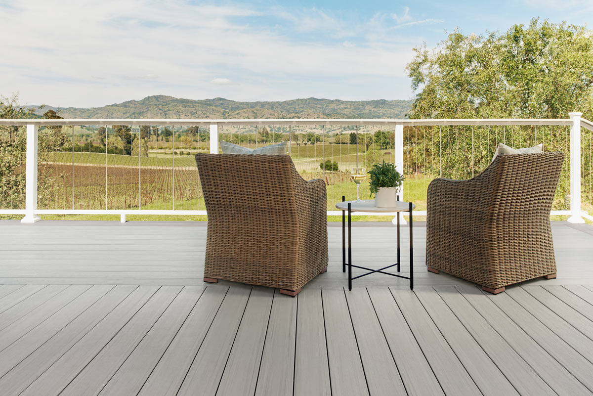 A patio, facing the mountains with lush greenery and vineyards, decorated with with wicker chairs and a short round table with a green fern plant. 