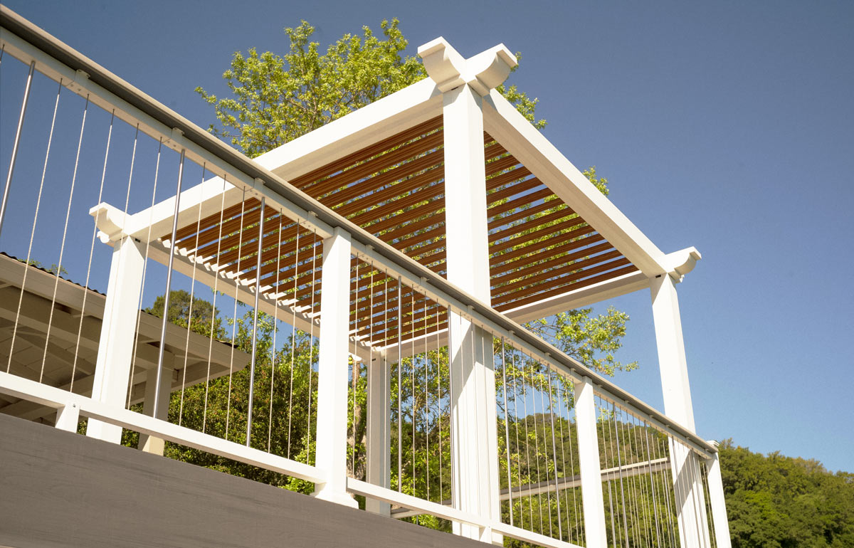 A view of the white pergola with a red planking roof from below. The pergola stands out agains the blue sky and the green tree.