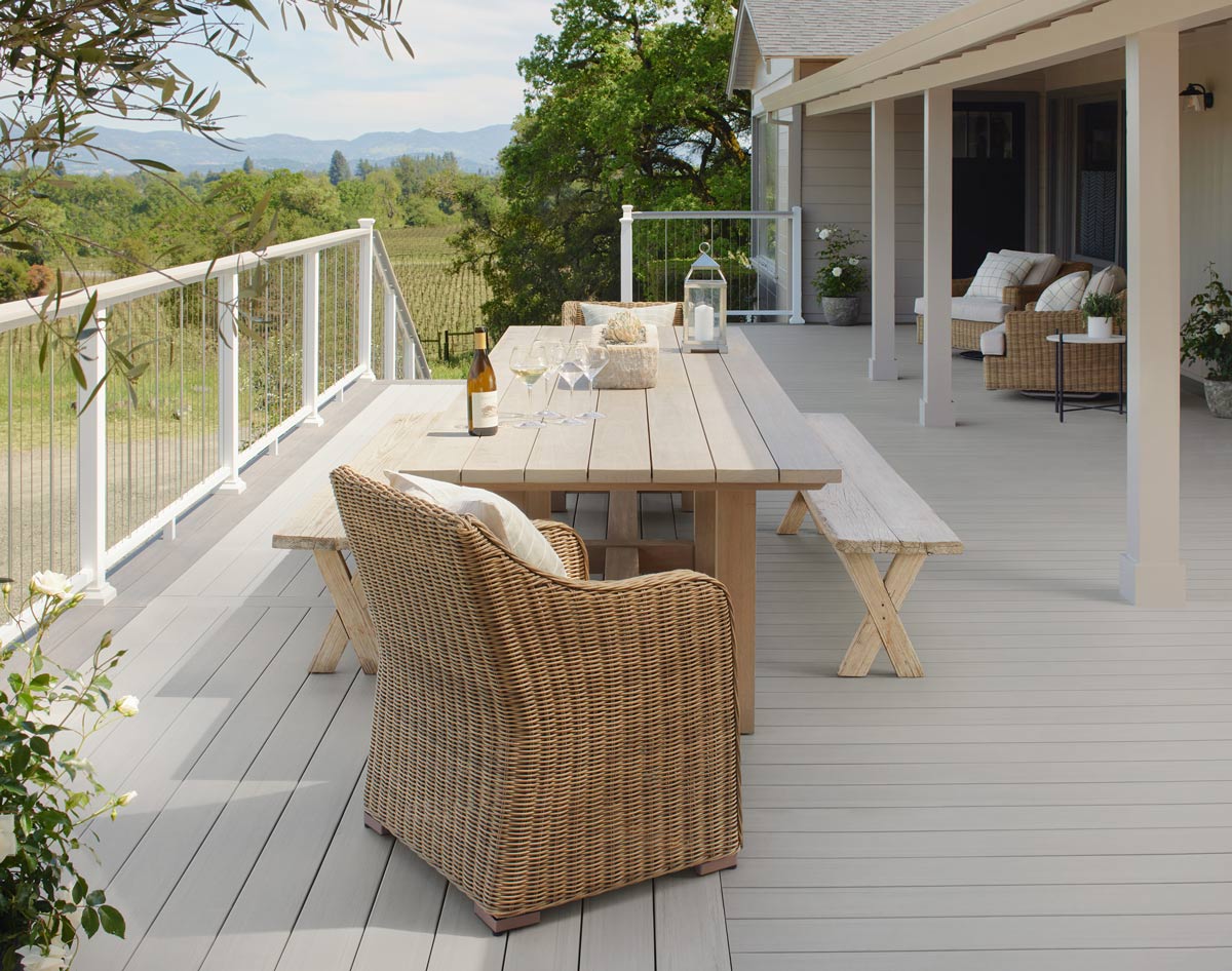 A closer view of the white railing with the tan table. The table is decorated with a one half filled goblet of wine and three empty glasses next to a open bottle of wine. A view of the winery is visible.