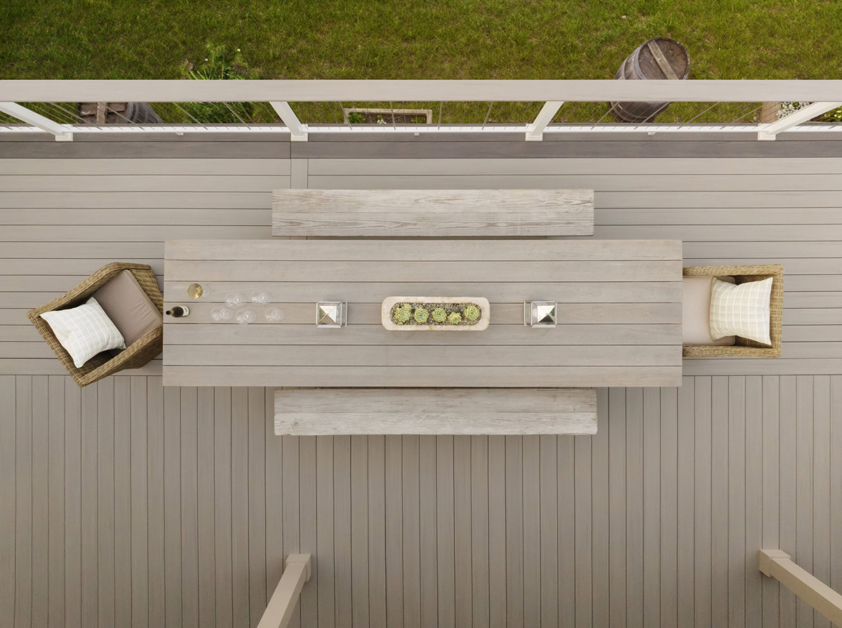 An over head view of the table with an arrangement of wine goblets and an open bottle of wine, lanterns with reflective lids, wicker chairs and benches. A detailed view of the brown decking and white drink rail. 