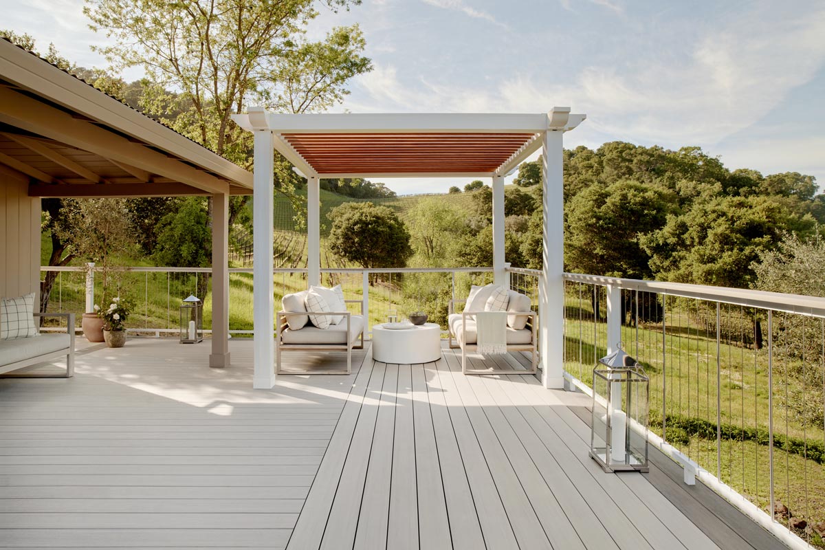 A pergola painted white and decorated with white accent pillows and future on a brown decking.