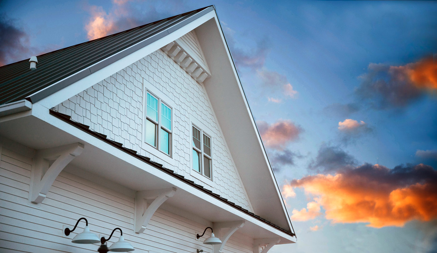 A view from below of white shingle siding and black roofing. Four windows looking up at the blue sky which is decorated with white, orange and dark purple clouds. 