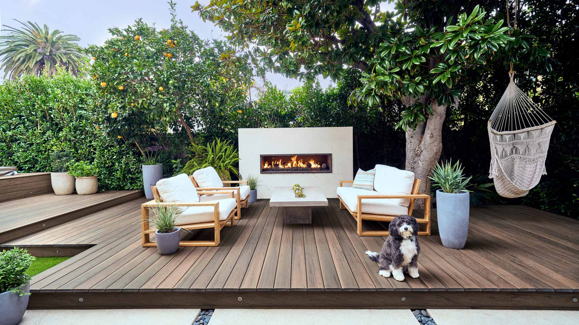 A backyard with a hammock, fireplace, furniture on dark brown Pecan decking with black and white dog in the foreground.