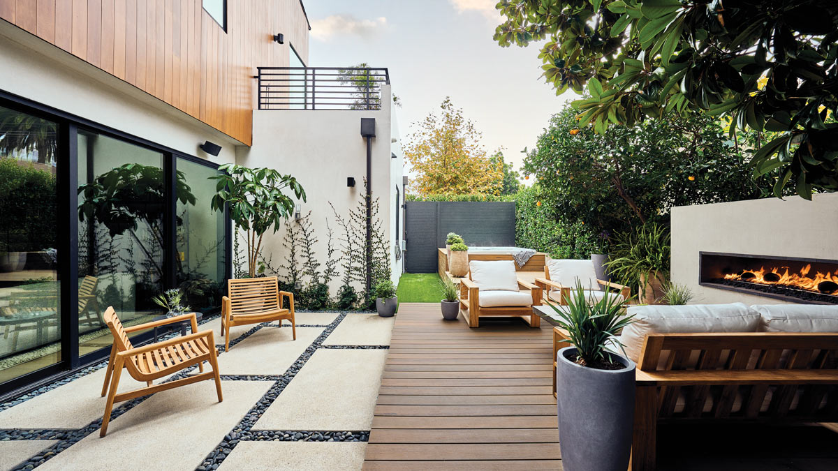 Backyard facing a dark metal second floor patio rail with small fruit trees on the right and fireplace across from two wooden chairs facing reflective floor to ceiling windows. 