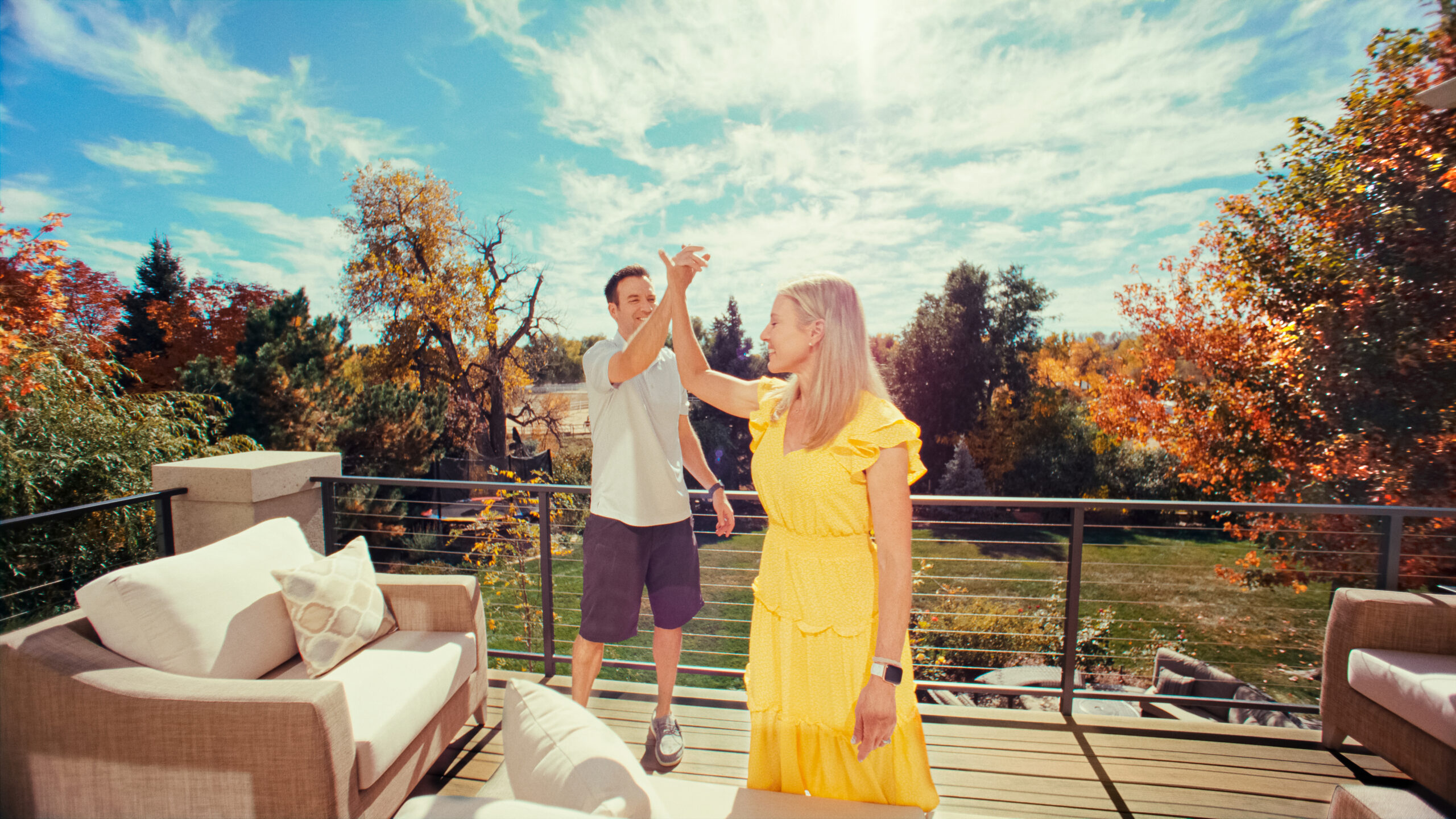 Jason and Kelly high-five on their deck as Kelly wears a yellow dress and Jason wears a shirt and dark gray knee-length shorts. The deck is decorated with an armchair with a patterned pillow. The couple stands against a backdrop of trees with green and orange leaves. The blue sky is littered with scattered white clouds above them.