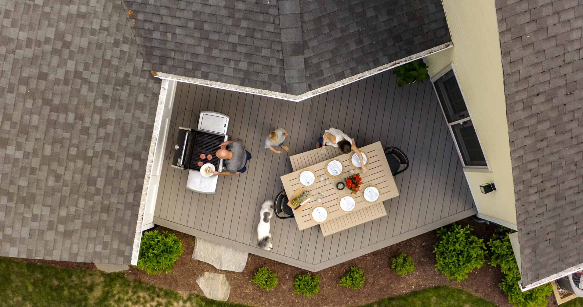 An aerial view of Deb and Rob's deck area that includes a grill and a tan table with plates and benches. Their white and brown dog circles the table as Rob places some table decor on the table.