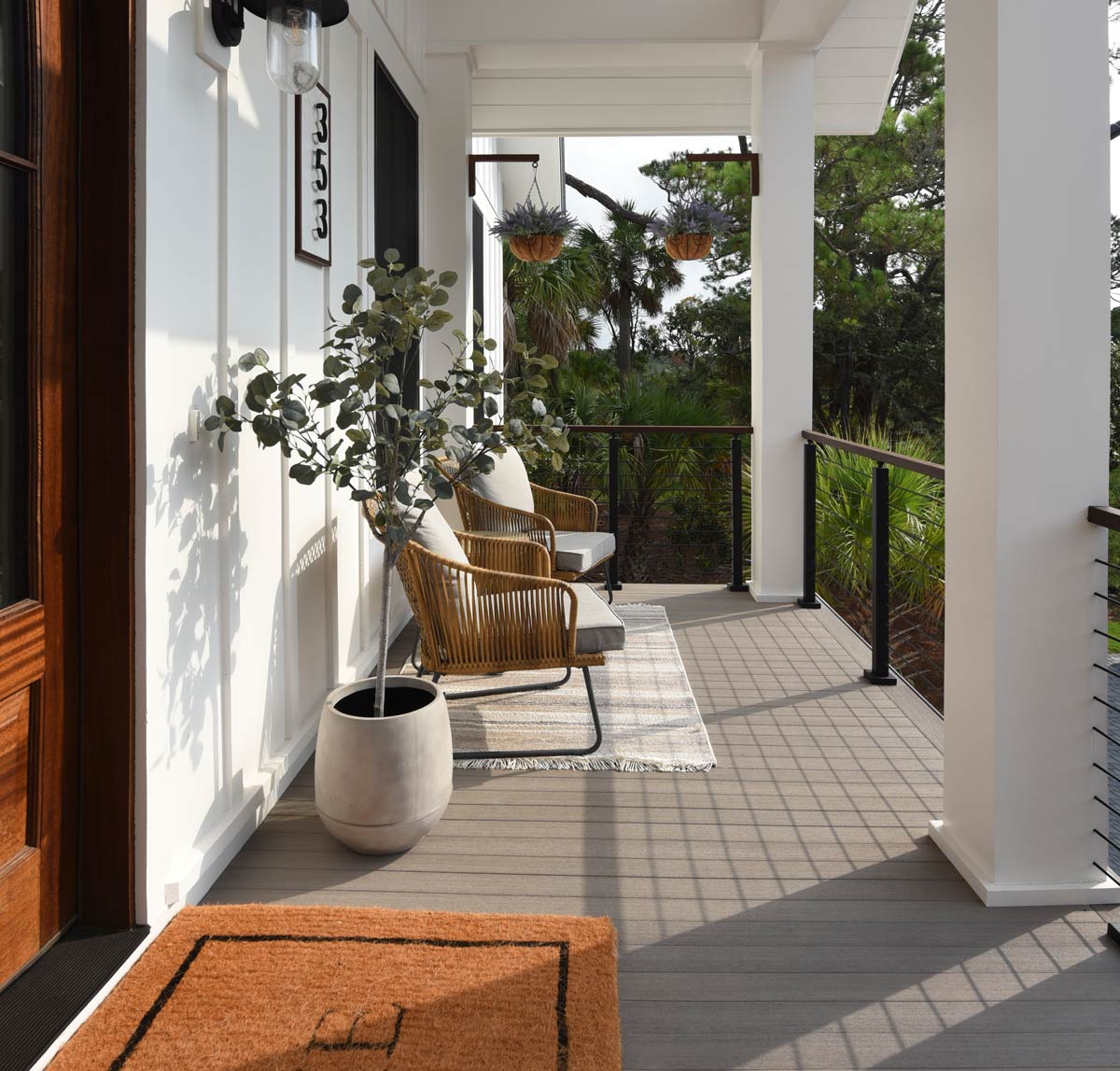 A front porch featuring a doormat, two wicker chairs with cushions, a potted plant, hanging plants, and a house number "353" plaque. The porch has white siding and overlooks a natural landscape.