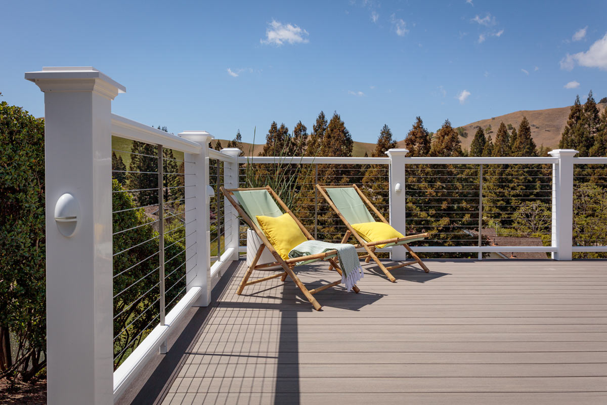 Two cabana chairs with mint green slings and yellow throw pillows sit in the corner of a composite deck on a sunny day. Classic Composite Series Premier Rail in White runs along the deck's perimeter and features a stainless steel cable rail infill. Beyond the railing are tall coniferous trees and beyond that, hills in the distance. The composite deck boards are Coastline from the TimberTech AZEK Vintage Collection. The railing is the Classic Composite Series Premier Rail in White.