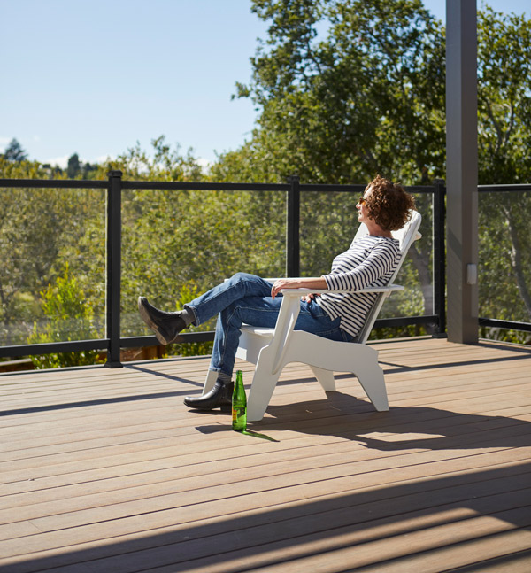 A woman relaxes in an adirondack chair and looks at the trees that are visible past the deck railing with glass infill