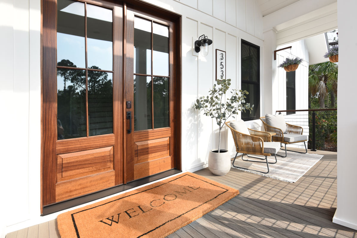 A welcoming front porch with a large wooden door, a "WELCOME" doormat, two chairs, a small table, and hanging plants. The dusty gray Coastline decking is from TimberTech's Porch Collection.