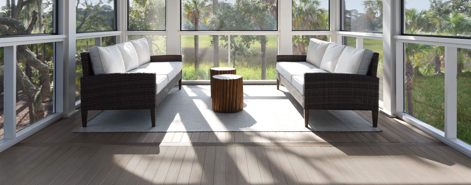 A bright, sunlit screened-in porch with medium gray decking in the Coastline color from the Porch Collection by TimberTech. The porch features two dark wicker sofas with white cushions facing each other. A small wooden stool is placed between them on a light-colored rug, while large floor-to-ceiling windows showcase an outdoor view of trees and greenery.