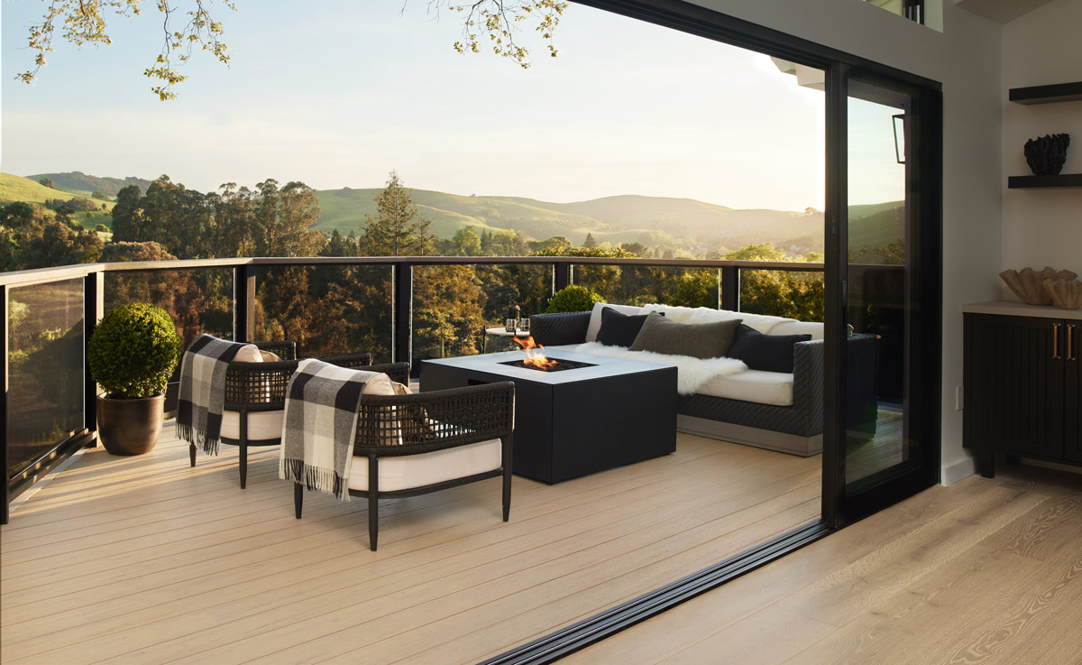 A balcony deck with Reclaimed Chestnut from the Reserve Collection with a drink rail with glass clear infills. The background looks out to a green of trees and green rolling hills.