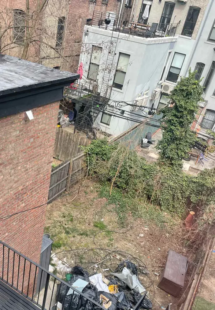 The overgrown Brooklyn backyard before the Real Simple and Decks R Us remodel. Garbage and piles of refuse are piled up in the backyard.  A view from a balcony on the backside of the brownstone which displays the neighboring buildings. 