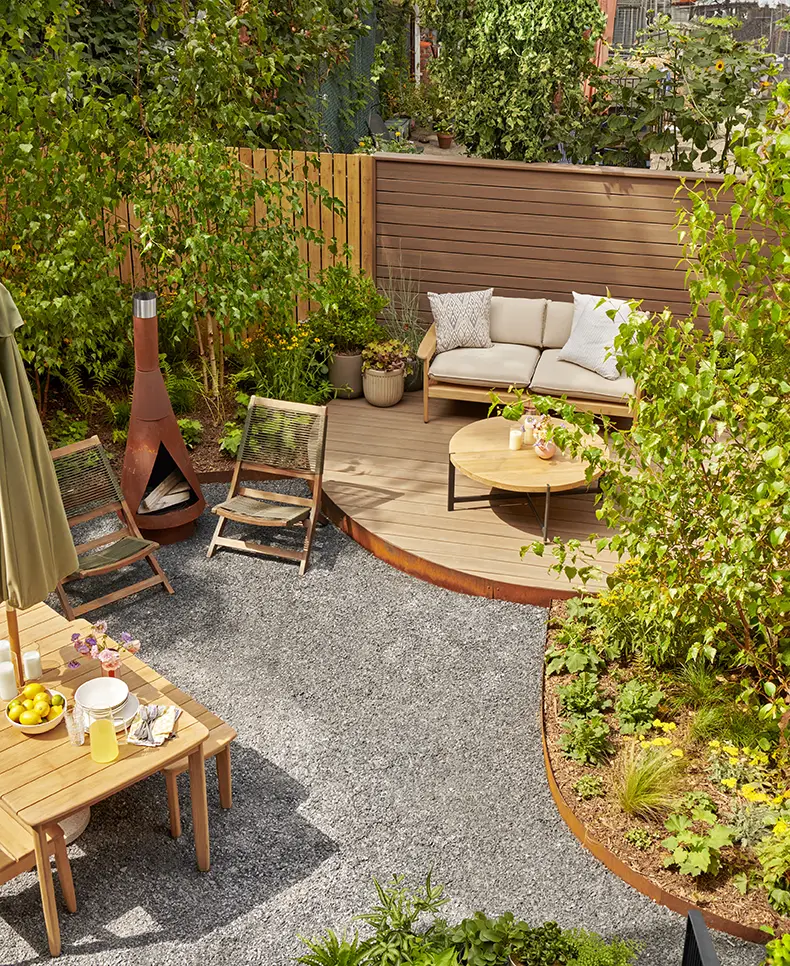 A sunny backyard patio decorated in various shades from the Vintage Collection in various shades of English Walnut. The ground is blanketed in dark gray rocks. The patio is decorated with bushes in variety of stages of growth. The tables are decorated with plates, bowls and glasses of lemonade. The landscape design. was created by Brooklyn Grange. 