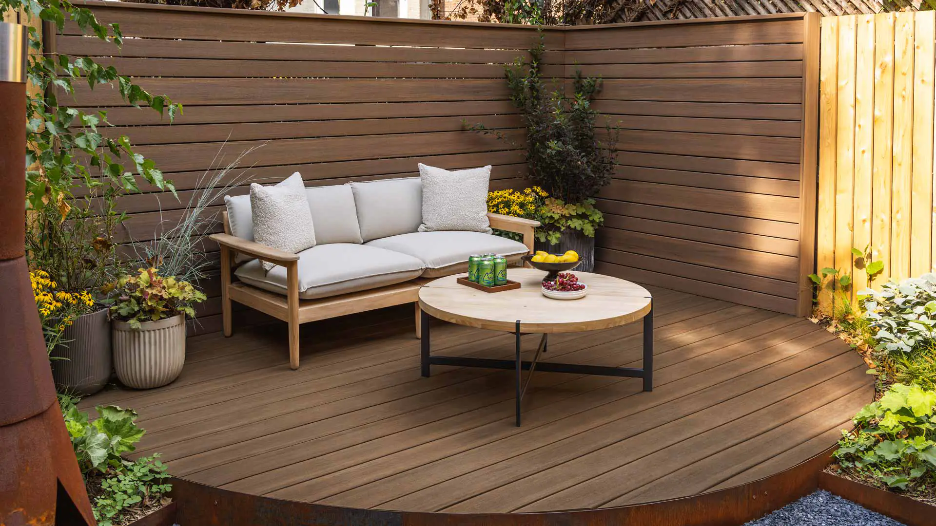 A view of backyard couch on English Walnut decking and walls from the Vintage Collection. There is a patio couch with beige pillows and light brown framing. The deck is adorned with green planted in pots and planted in the ground. 