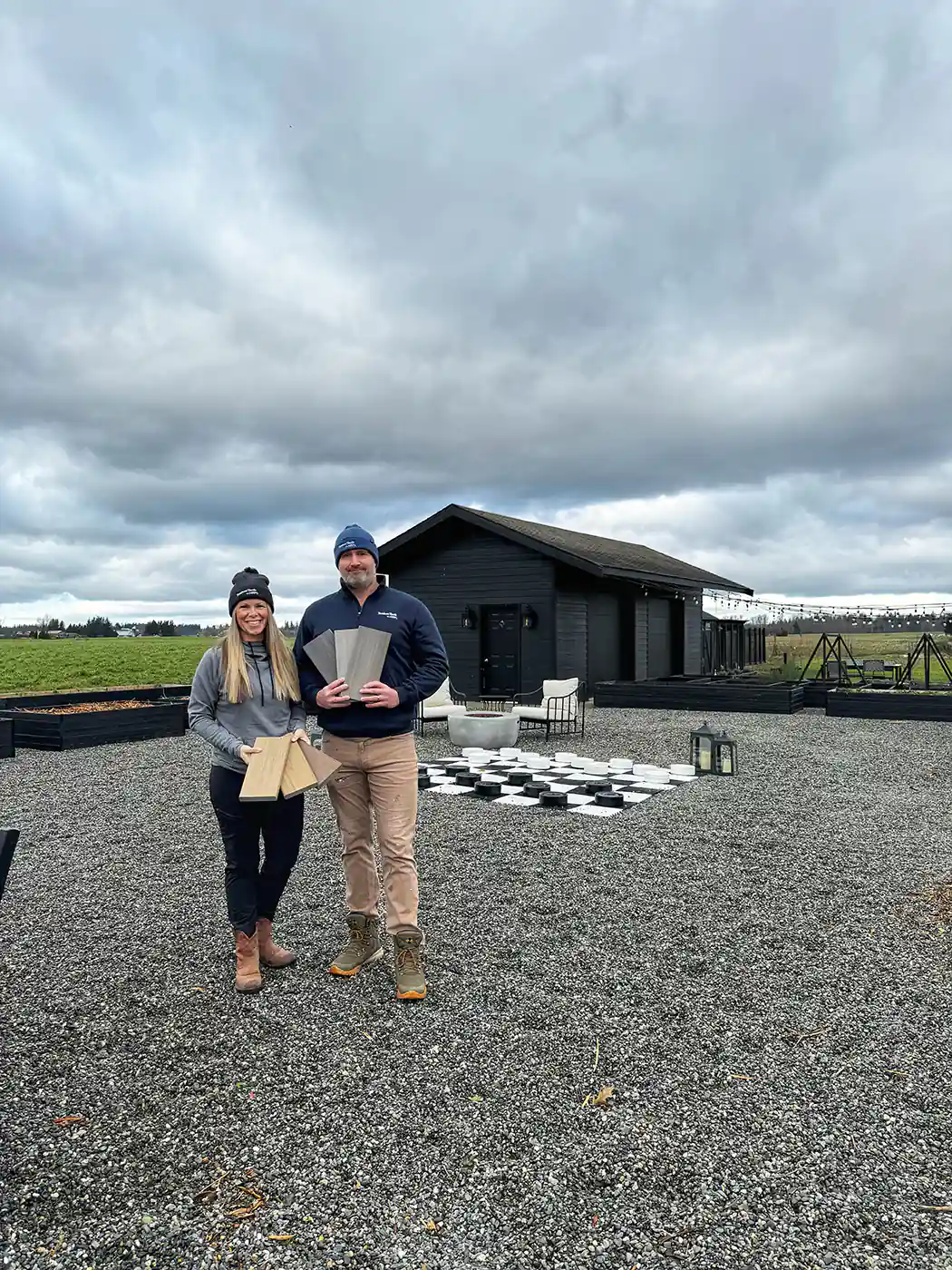 Sarah Wagner and her husband James Wagner holding their Timbertech swatches, in front of decking black and white chessboard under an overcast sky with white fluffy clouds. This was before the full deck was built. 