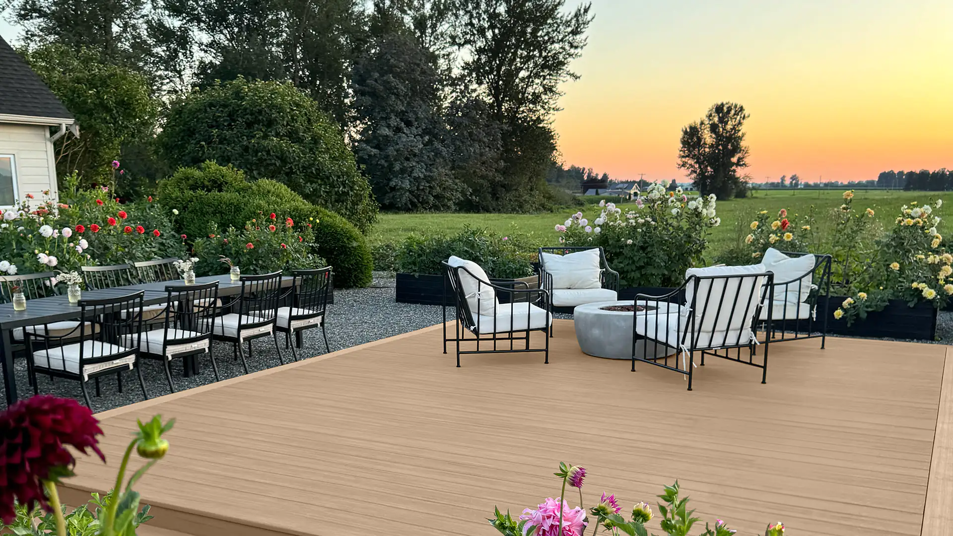 A set of black iron wrought chairs with white pillows surround a gray unlit fire pit. This arrangement sits atop a light tan deck in shade of Weathered Teak from the Vintage Collection. A black table is on the left side of the image with iron wrought dining set with white pillows and sits atop a gray rock filler. The deck is surrounded in an assortment of red, white and yellow flowers and green bushes.